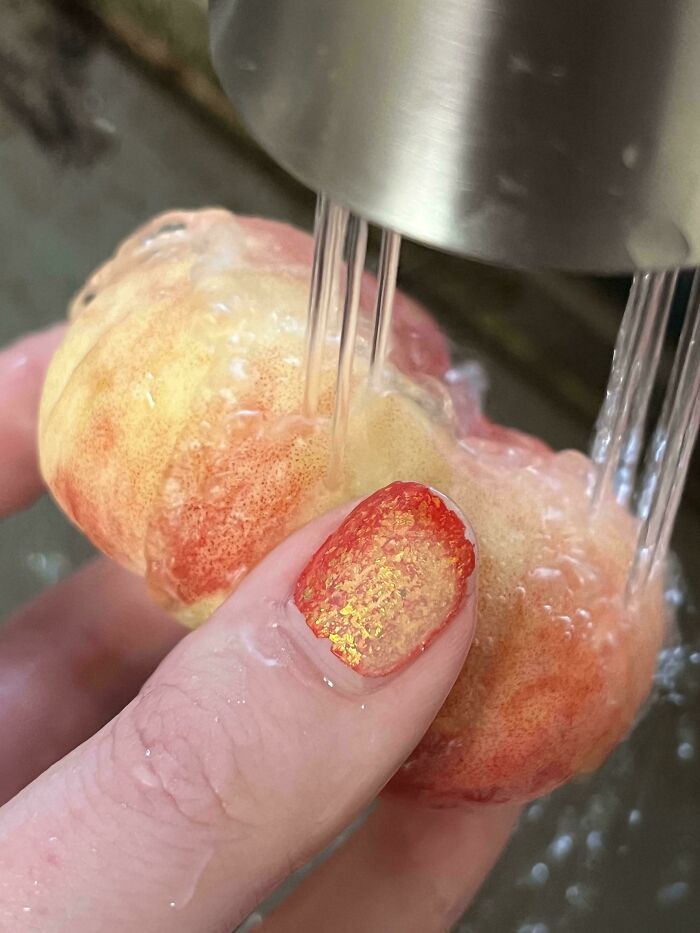 A hand with shimmery orange nail polish holding a small, light-colored fruit under running water, demonstrating satisfying paint matches.