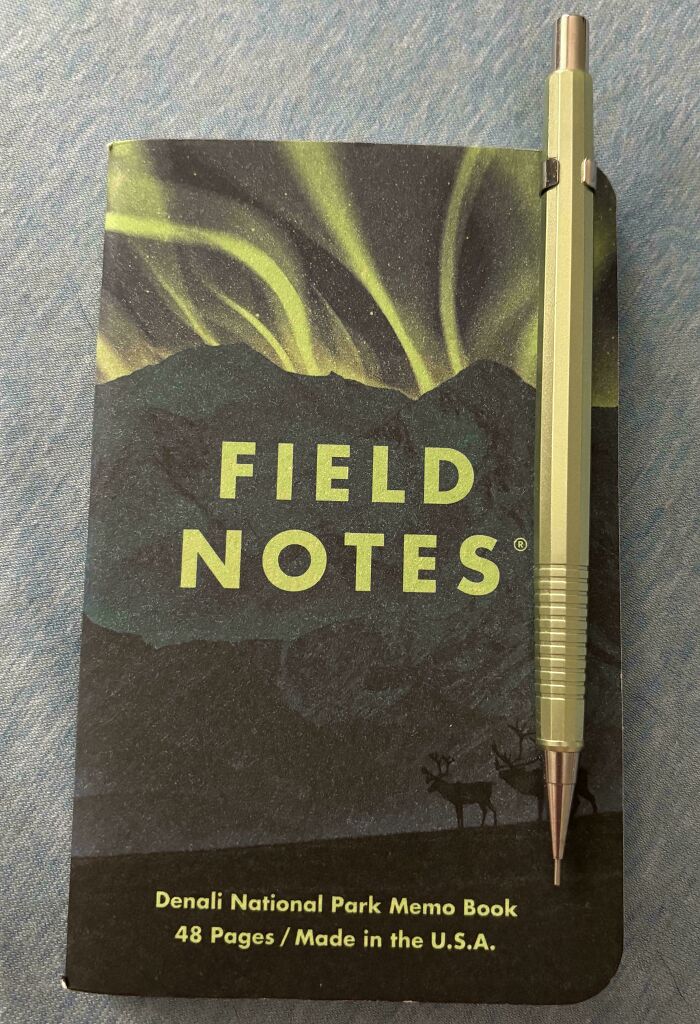 A Field Notes memo book with a green aurora borealis design, next to a matching green pencil, displaying satisfying paint matches.