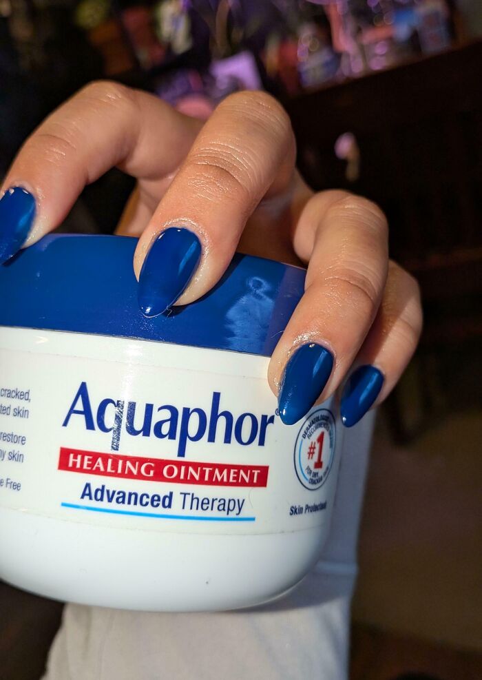 A person with shiny blue nails holds a jar of Aquaphor Healing Ointment, the nail color perfectly matching the jar's blue label, an example of satisfying paint matches.