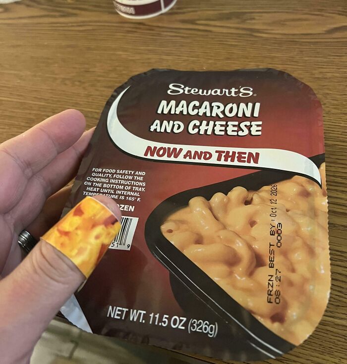 A hand holding a Stewart's Macaroni and Cheese package, with a bandage on a finger that is a perfect paint match to the macaroni.