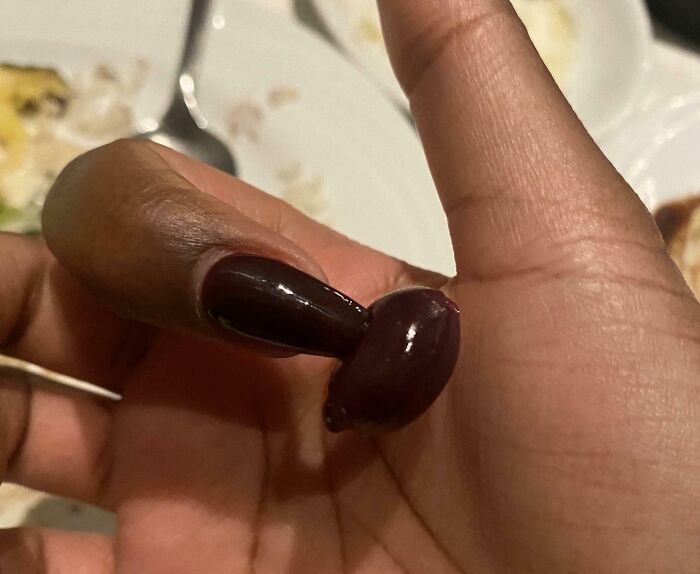 A hand with dark, glossy fingernail polish holding a dark olive, showing a satisfying paint match between the nail and the olive.