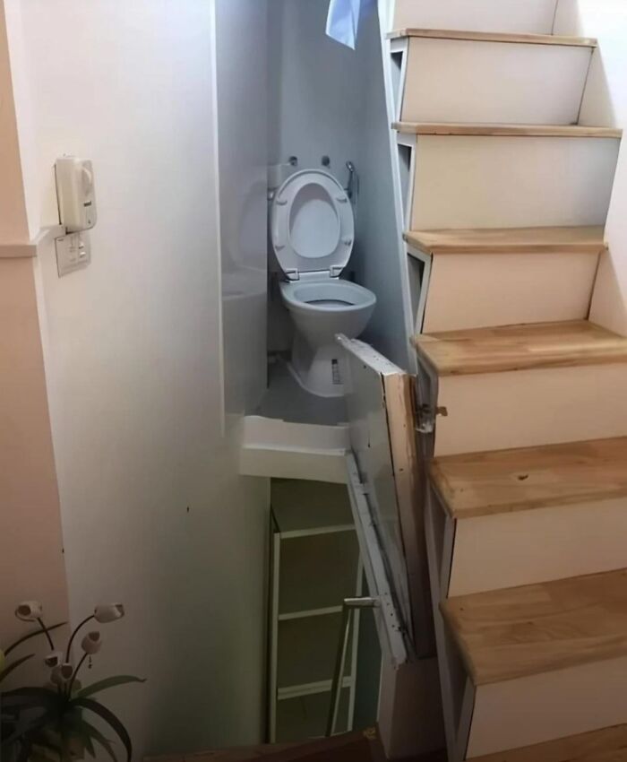 A terribly designed bathroom with a toilet awkwardly placed under a low staircase, next to a small storage space.