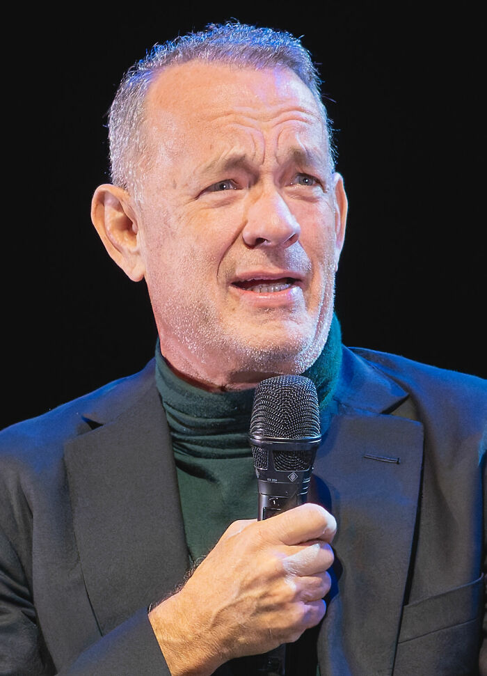 Tom Hanks, renowned actor, speaking into a microphone, sharing insights about his bio and career highlights.