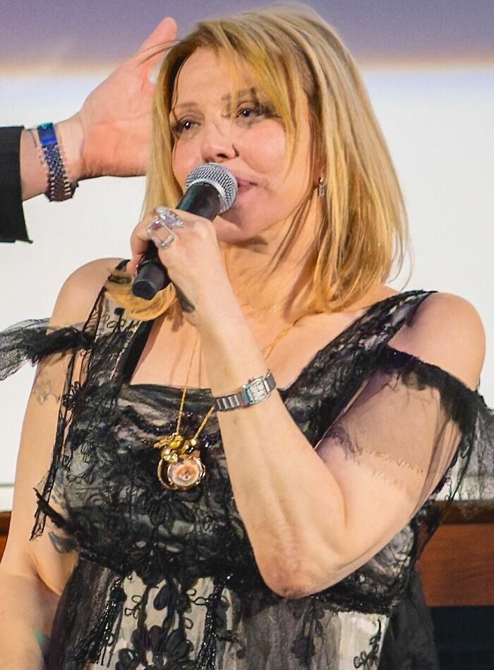 Courtney Love on stage, performing during her career. She is holding a microphone and wearing a black lace top.
