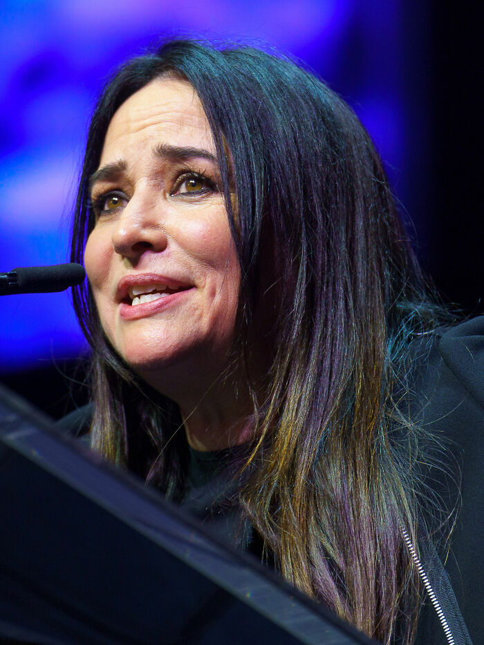 Pamela Adlon speaks at a microphone, highlighting her career. Her long, dark hair has subtle streaks of color. This image focuses on her profile.