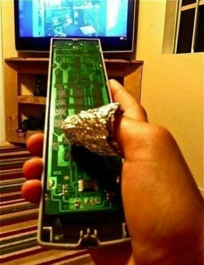 A hand holding a TV remote with a missing back cover, showing foil used as a wild fix for the battery compartment.