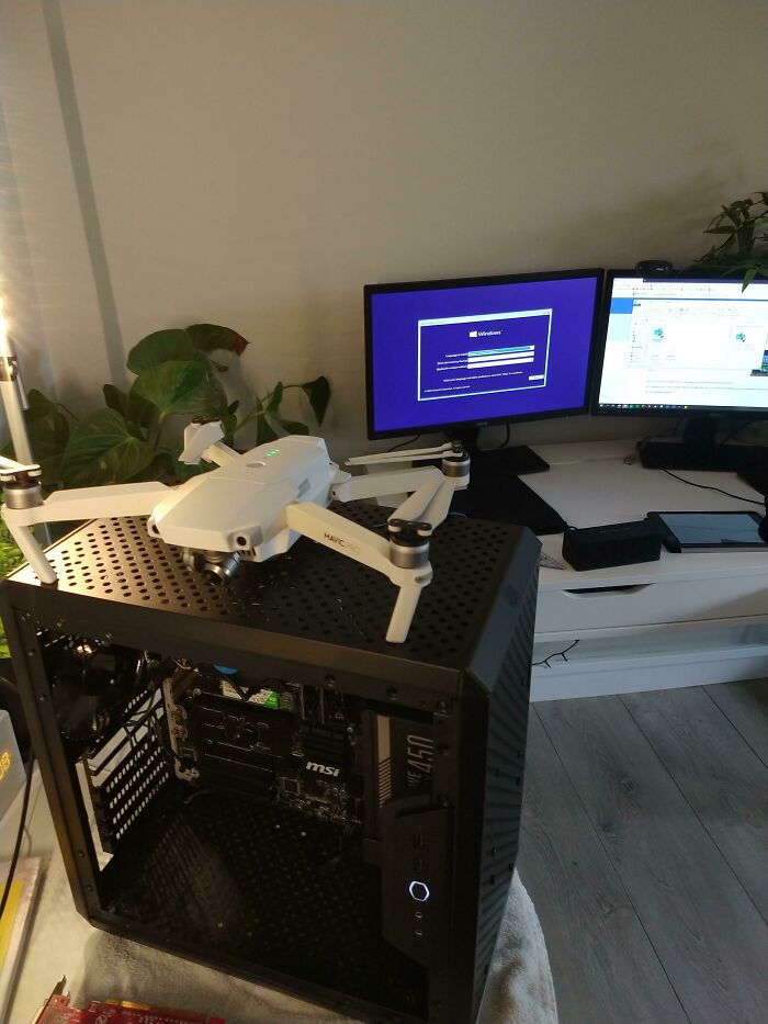 A drone sits on top of a PC case, serving as an unhinged way to fix something on a desk with monitors.