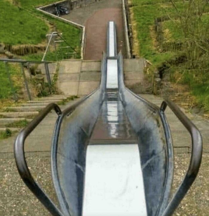 A long, winding metal slide in a park with stairs and grassy hills, a common sight that brings back good memories of the 80s.