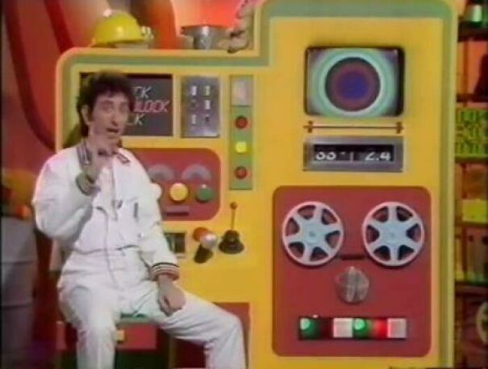 A man in a white jumpsuit gestures beside a large, colorful machine with reels, bringing back good memories of life in the 80s.