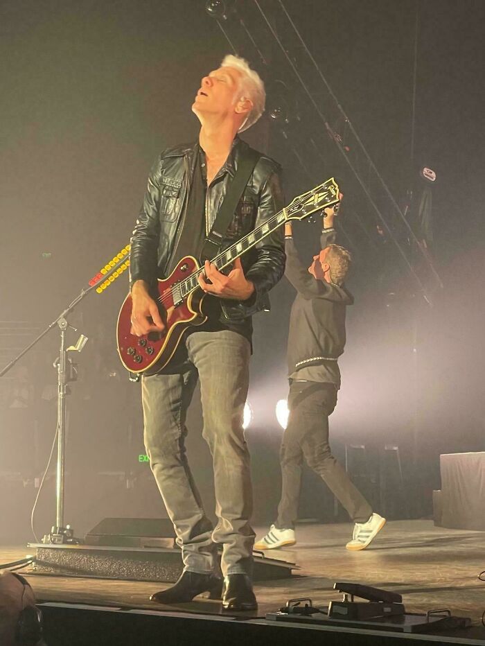 A man playing a guitar on stage with a person behind him holding an object creating an optical illusion of a giant hand. Look twice.