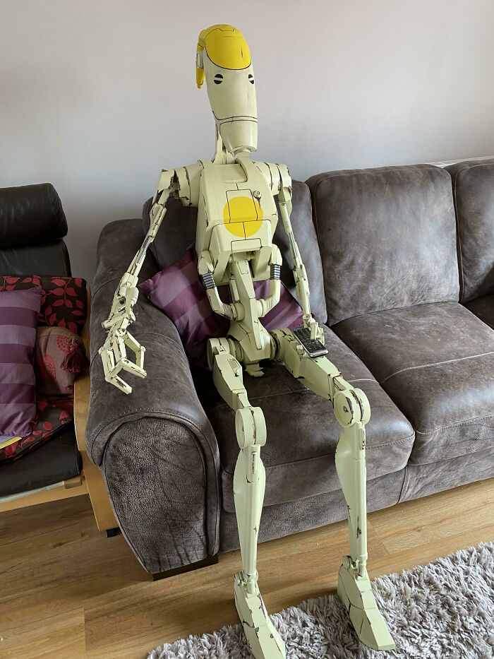 A large, detailed 3D printing of a Star Wars battle droid sitting on a brown couch holding a remote, showing amazing creations.