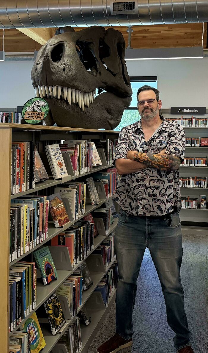 Man standing next to a large 3D printing dinosaur skull in a library, showing amazing creations.