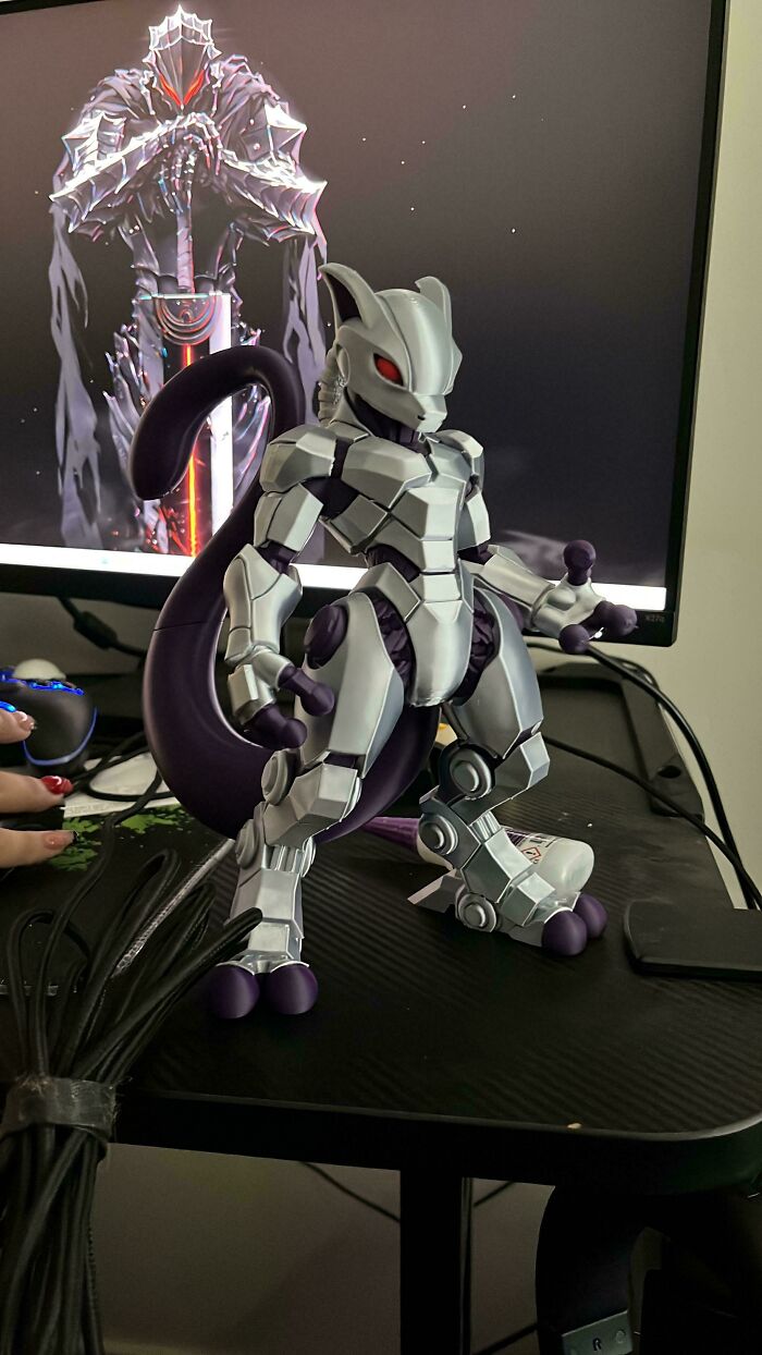 An incredible 3D printing creation of a metallic Mewtwo stands on a desk in front of a computer monitor.