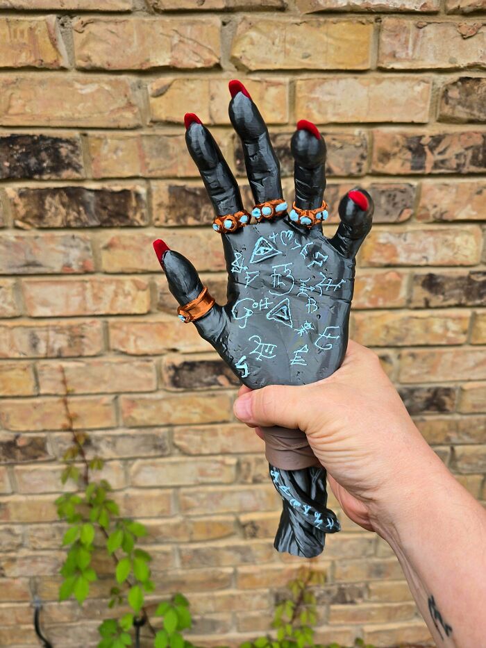 A person holds up an amazing 3D printing creation of a black, witch-like hand with red nails and rings, against a brick wall.