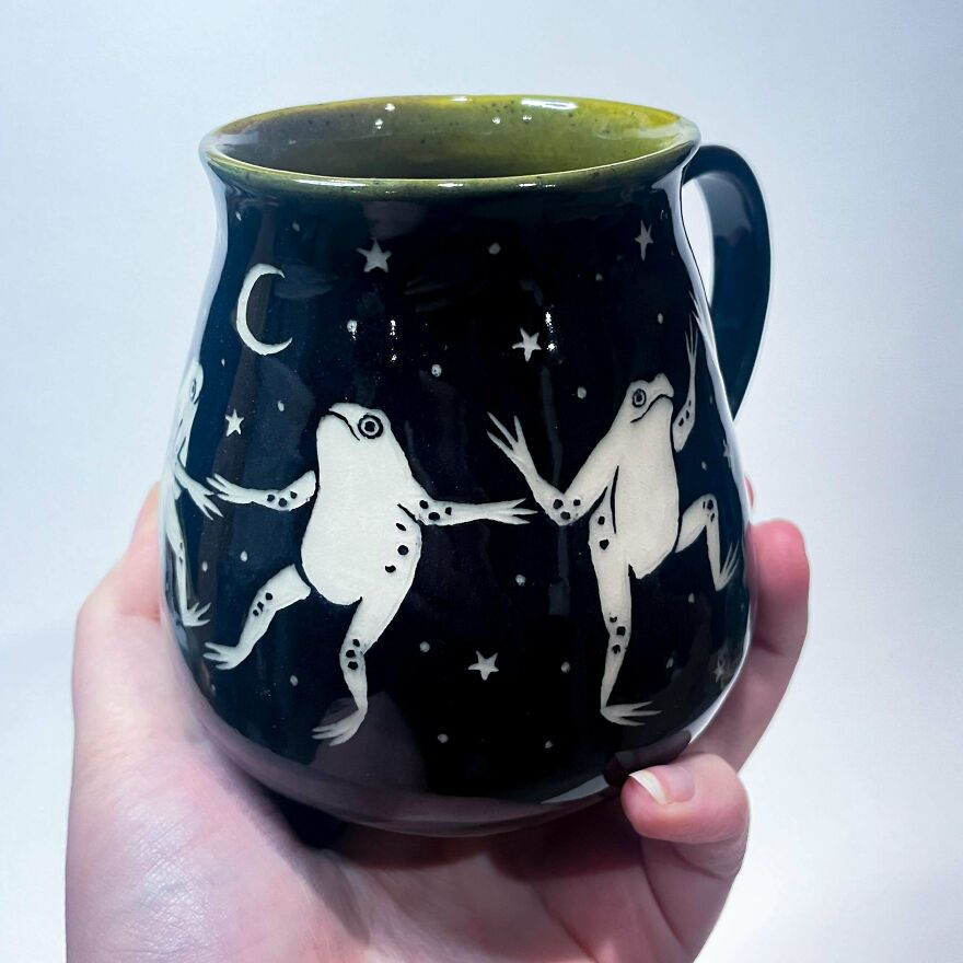 A hand holding a dark mug with whimsical crafts depicting white frogs dancing under a crescent moon and stars.