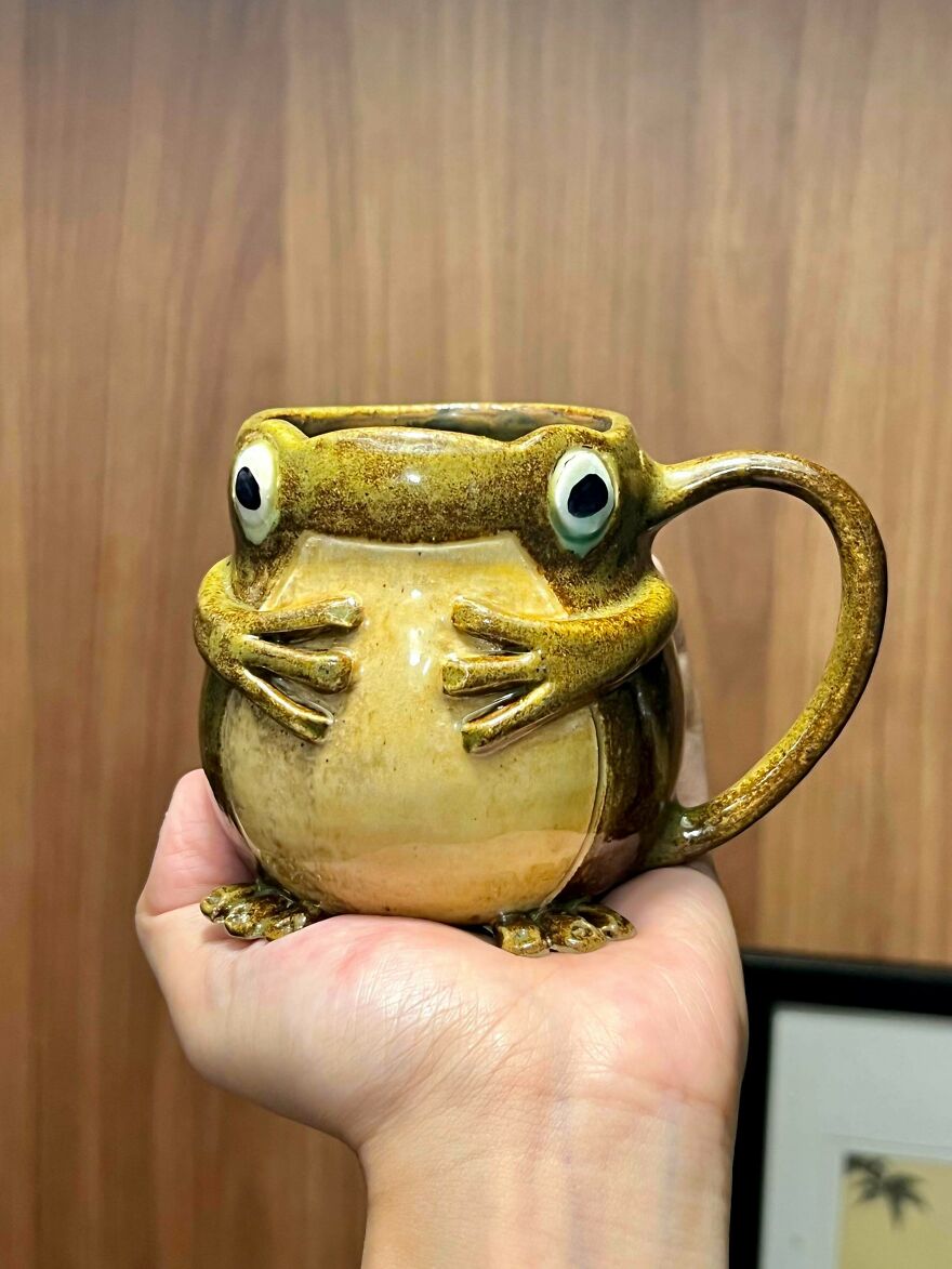 A hand holding a whimsical crafts mug shaped like a round, green and brown frog with big eyes.