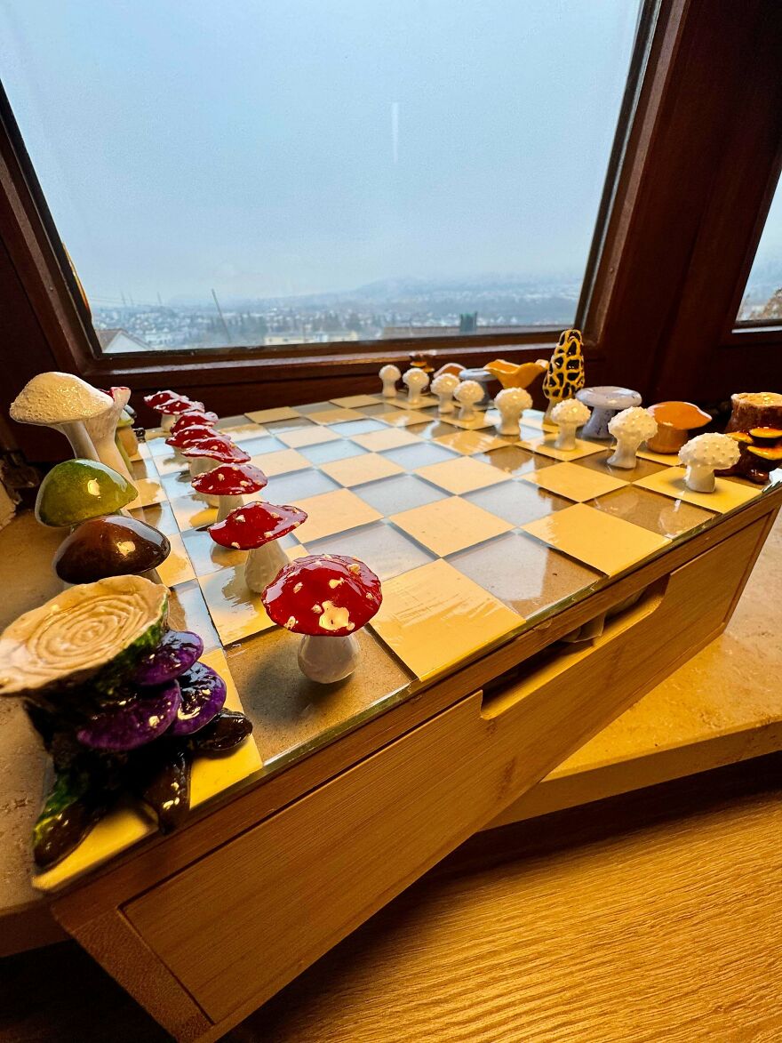 A whimsical craft chessboard featuring mushroom and tree stump pieces, set against a window with a scenic view.