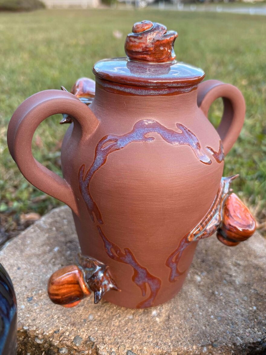 A whimsical craft of a reddish-brown pottery urn with two handles, decorated with textured, shiny orange and blue designs.