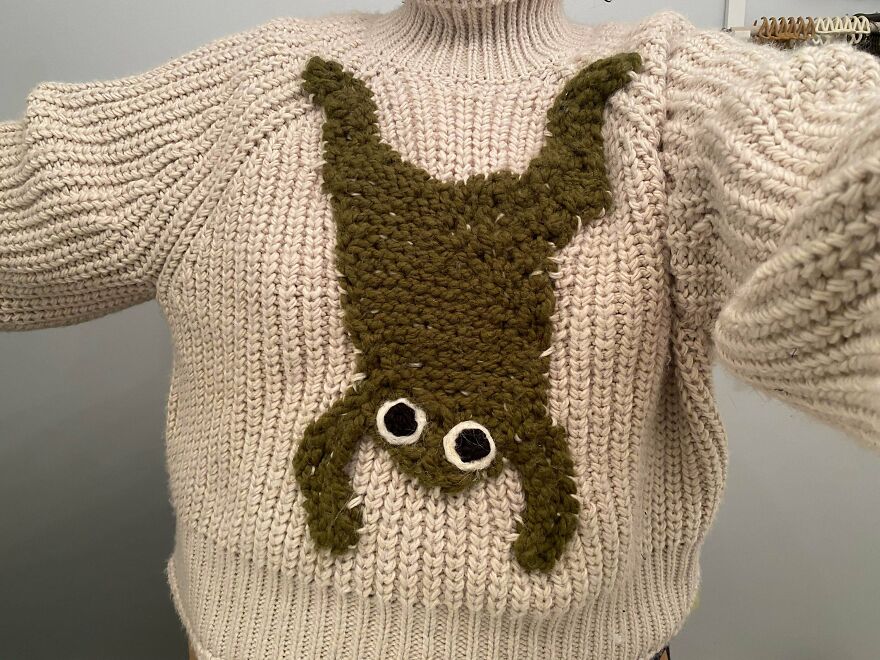 A person wearing a cream-colored, ribbed sweater with a large, olive-green knitted frog whimsical craft on the front.