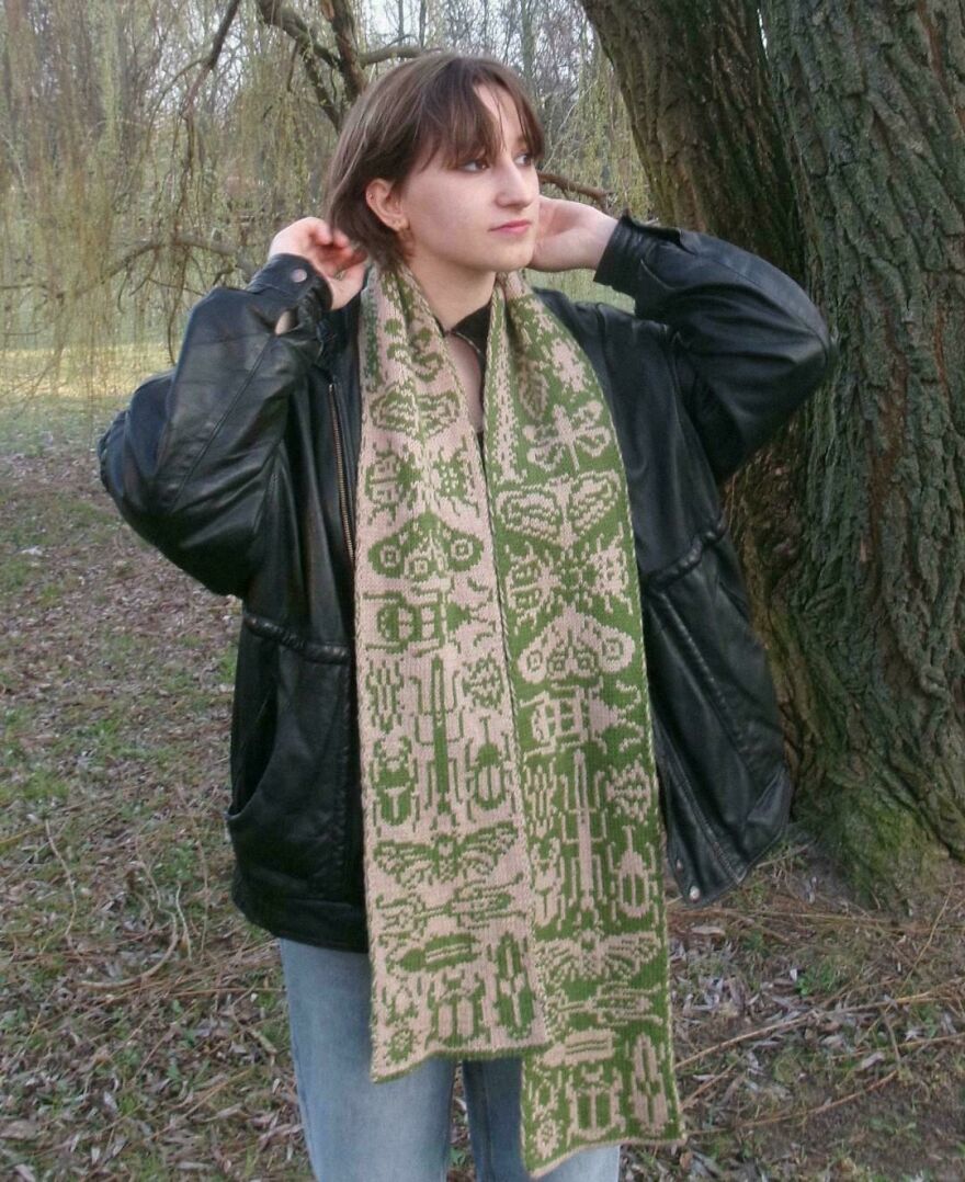 A person outside wearing a green and beige knitted scarf, showcasing a whimsical craft design against a tree.