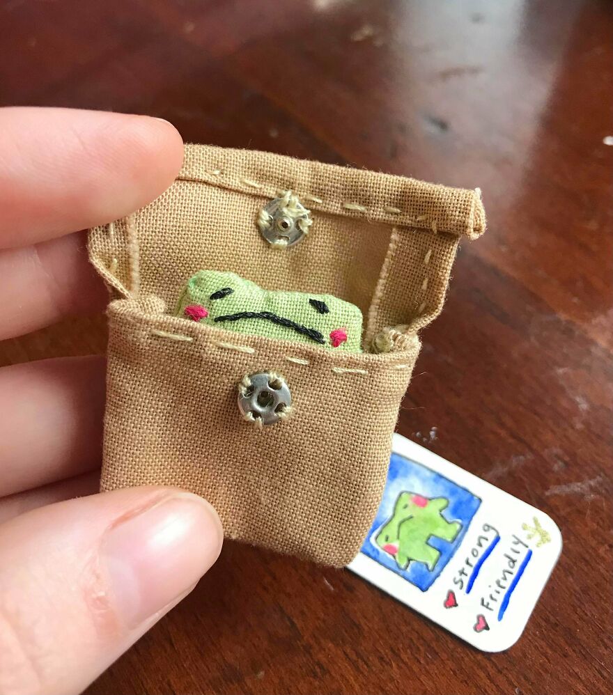 A tiny green whimsical craft frog tucked into a miniature fabric pouch with a tag reading strong and friendly.
