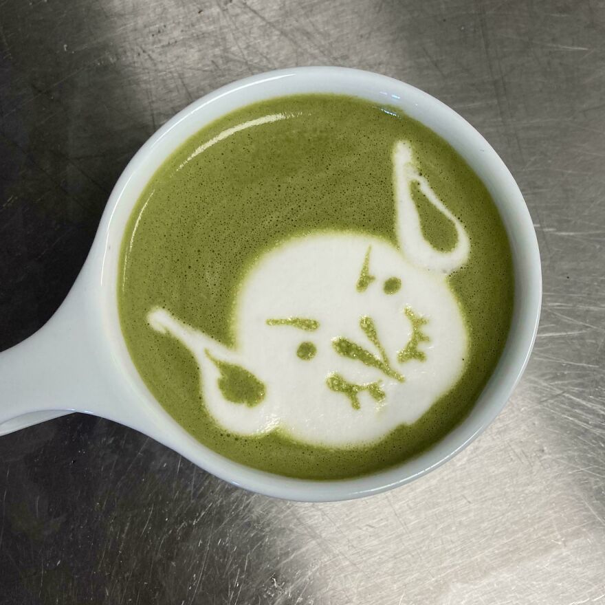 A cup of green matcha latte with whimsical craft foam art depicting a cartoonish angry face with pointed ears.