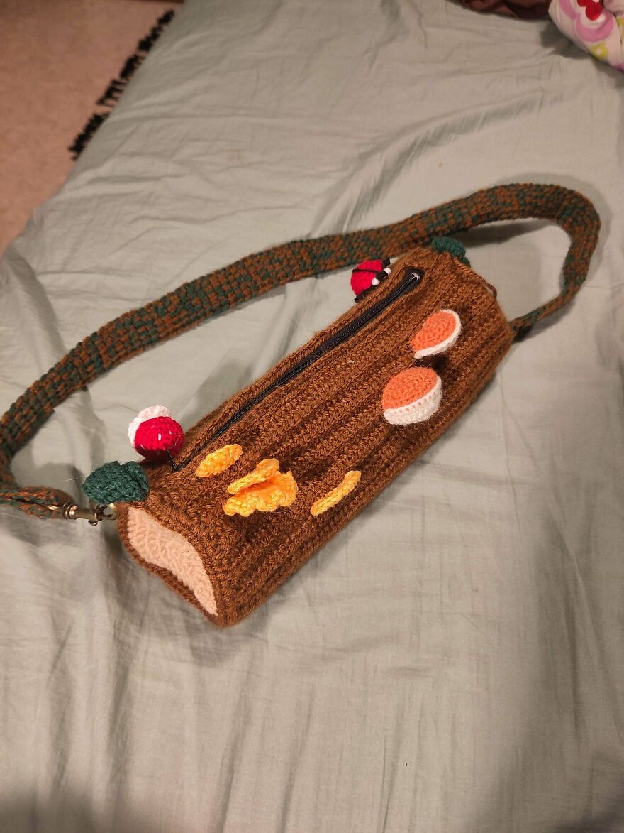 A whimsical craft crocheted log-shaped bag with a green strap, adorned with crocheted mushrooms and moss.