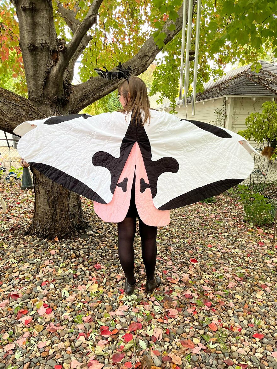 A person from behind wearing a white and black moth-wing cape, a magical whimsical craft, in a leafy yard.