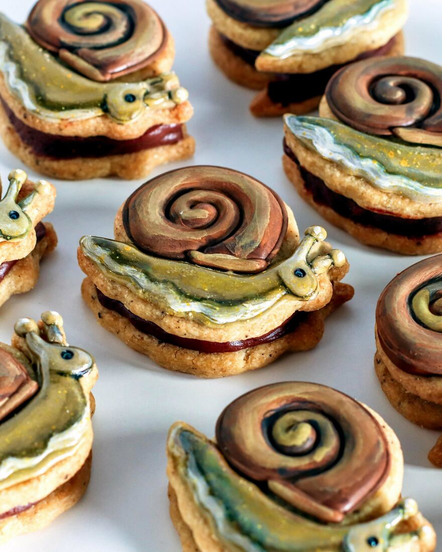 A close-up of several snail-shaped whimsical crafts cookies with detailed icing, some filled with red jam, on a white surface.