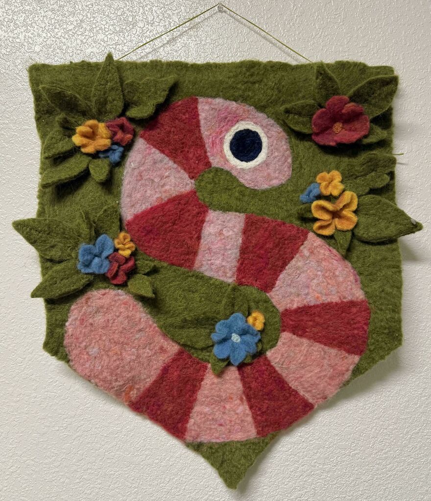 A felt whimsical crafts wall hanging depicting a red and pink striped snake-like creature surrounded by green leaves and colorful felt flowers.