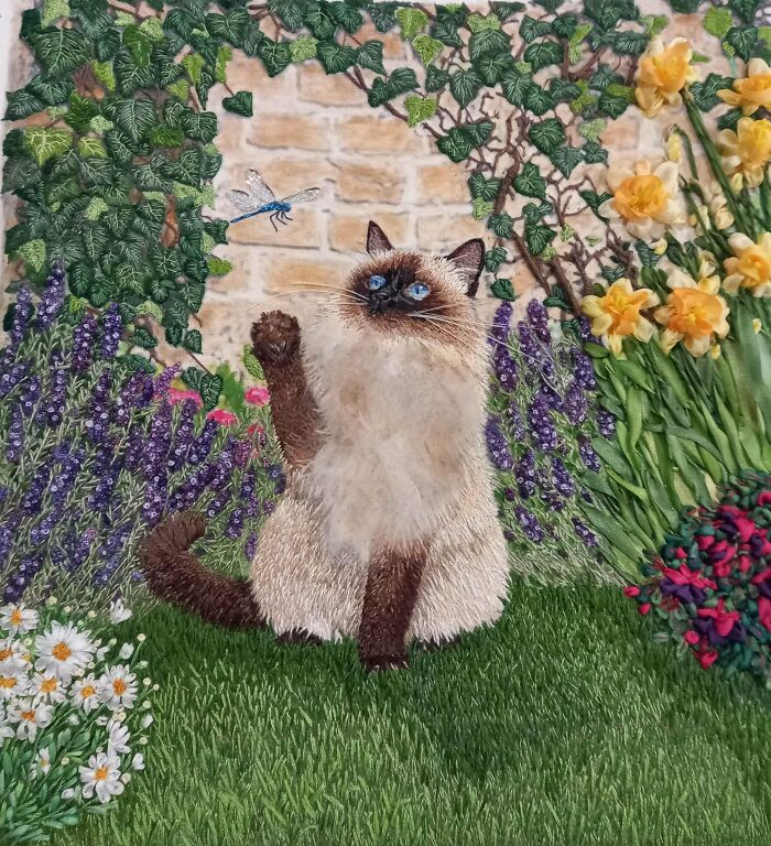 Detailed embroidery project featuring a fluffy Siamese cat looking at a blue dragonfly in a garden