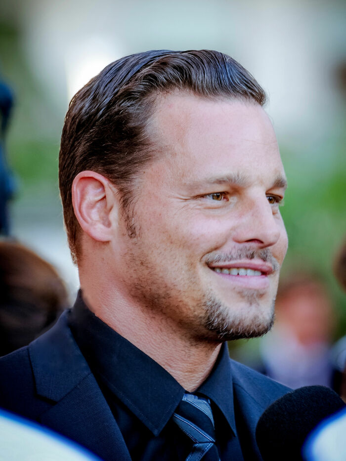A close-up of Justin Chambers smiling, wearing a dark suit and striped tie, showcasing his bio and career highlights.