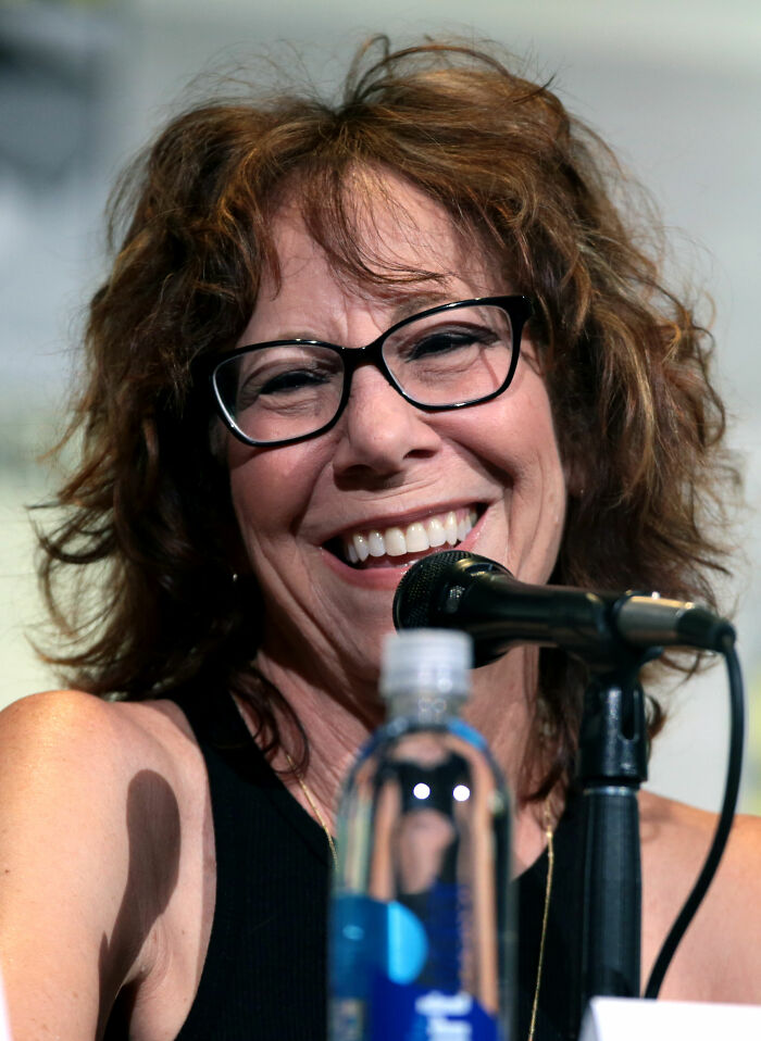 Mindy Sterling smiles broadly, wearing glasses, with a microphone and water bottle in the foreground, highlighting her career.