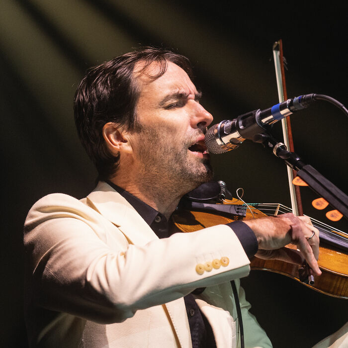 Andrew Bird performing live, singing into a microphone and playing the violin with intensity on stage.