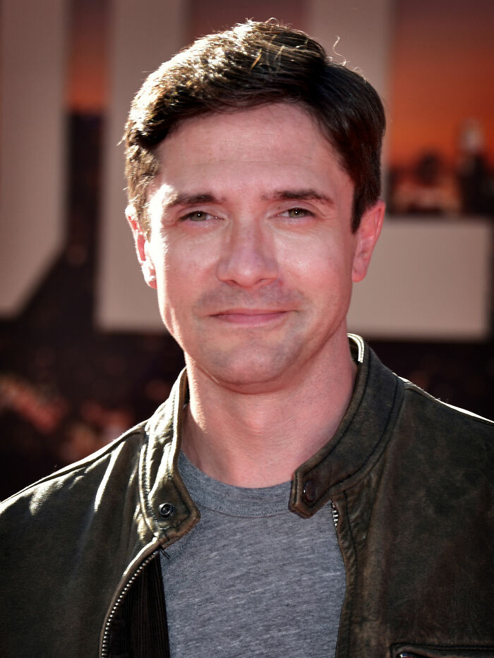 Topher Grace, known for his career highlights, smiles subtly while wearing a dark jacket.