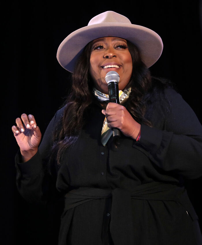 Loni Love, comedian, speaks into a microphone on stage, wearing a black dress, scarf, and hat.