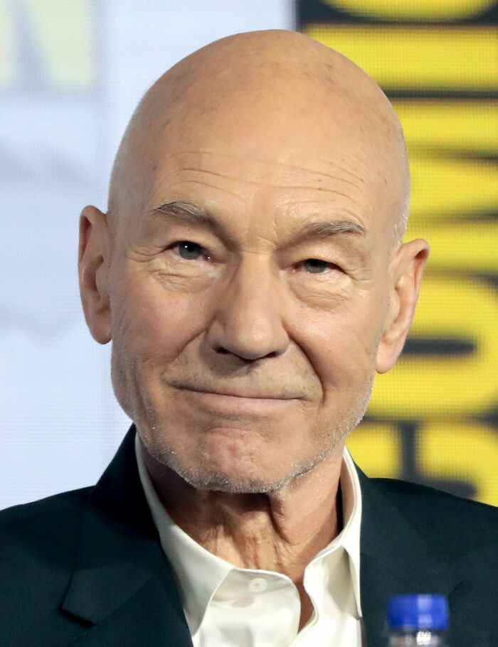 Patrick Stewart closeup, showing his bald head, white shirt, and dark jacket. Capturing his bio and career highlights.