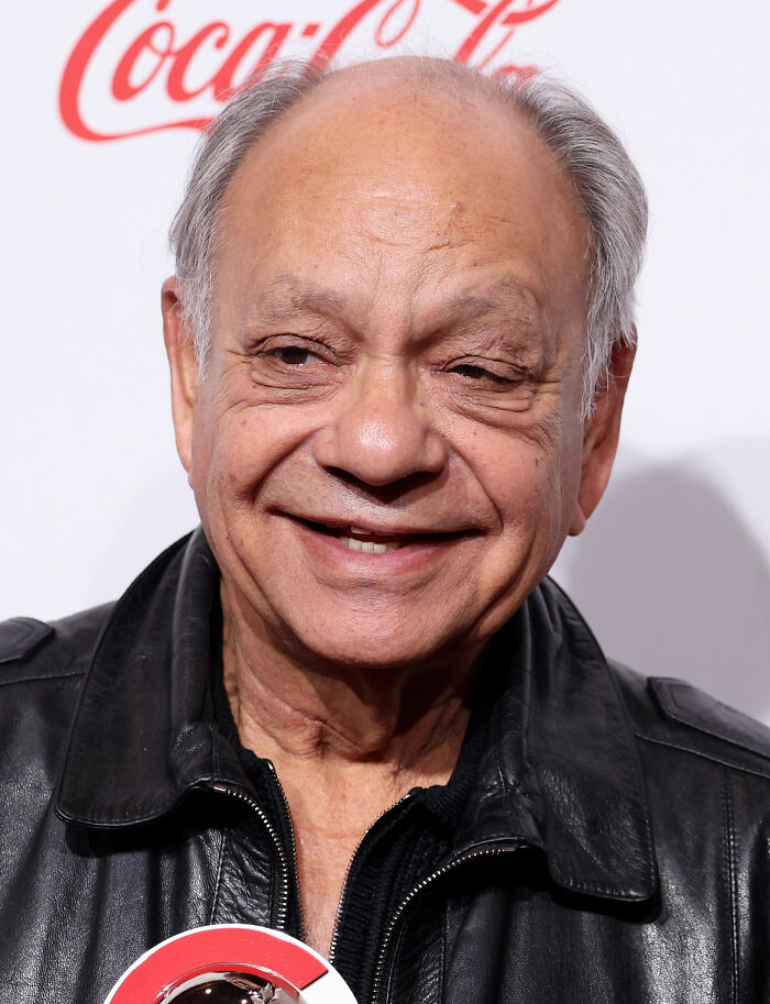 Cheech Marin smiling, wearing a black leather jacket, with a faint red Coca-Cola logo in the background. Bio and Career Highlights.