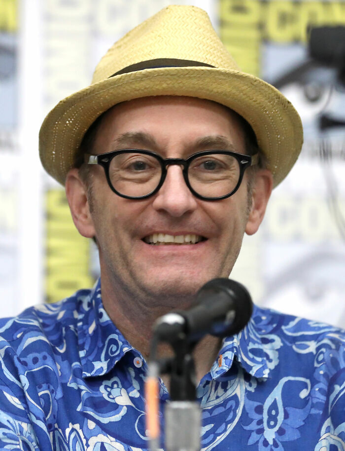 Tom Kenny, bio and career highlights of the voice actor, wearing a straw hat and glasses, smiling into a microphone.