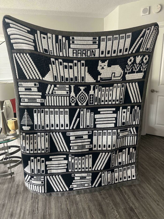 A large, detailed DIY crochet creation of a blanket designed like a bookshelf with books and a cat.