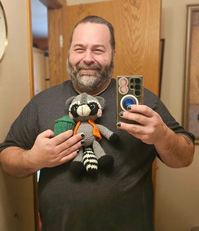 A man proudly holds a useful DIY crochet creation of a raccoon stuffed animal.