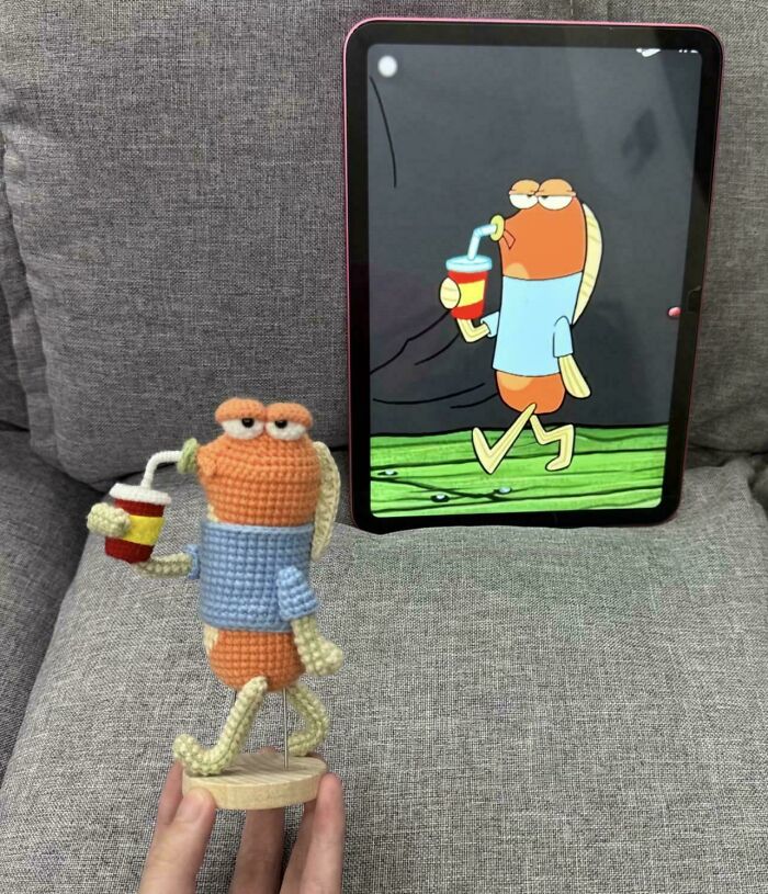 A person holding a crochet cartoon character of a fish drinking a soda, mirroring an image on a tablet, a fun DIY creation.