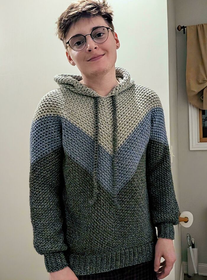 A person smiling, wearing a beautiful crochet hoodie with chevron patterns in blue, gray, and green colors, a useful DIY creation.