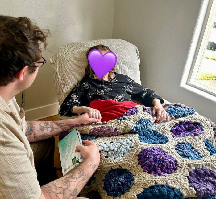 A person holding an elderly person's hand, who is covered with a multi-colored DIY crochet blanket, while reading a book.