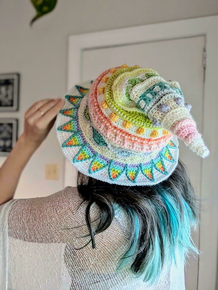 A person from behind wearing a colorful, pastel-striped DIY crochet wizard hat with a pointed top and a decorative brim.