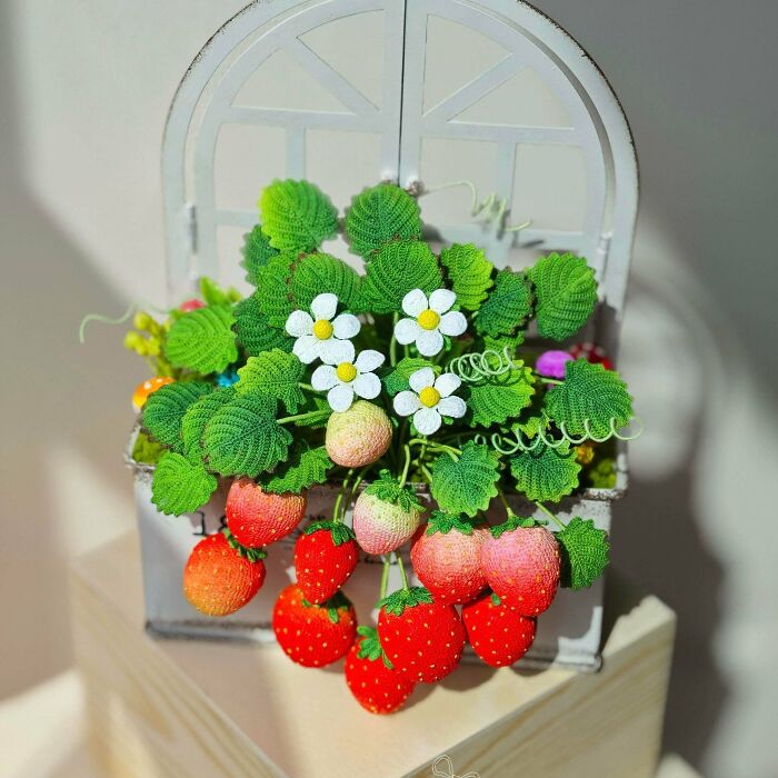 A DIY crochet creation of a miniature strawberry plant with red and green crochet strawberries and white flowers in a small white planter.