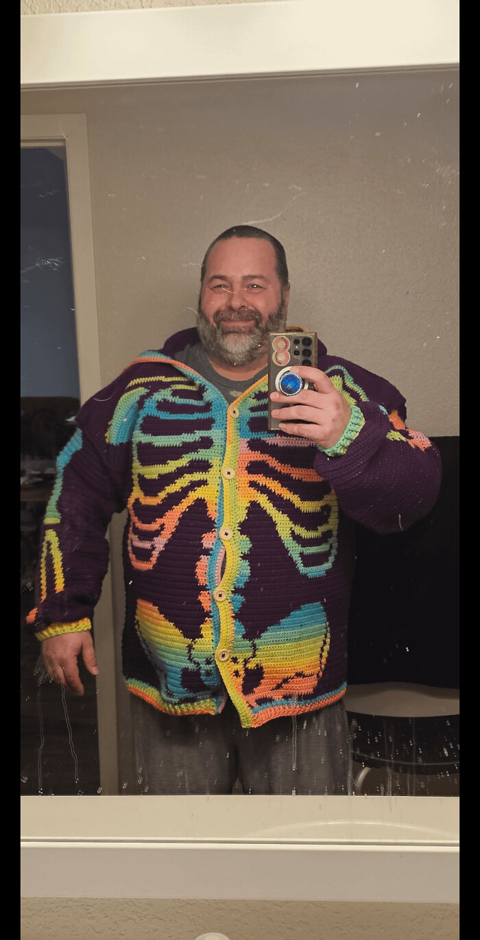 A man smiles in a mirror, showcasing his colorful skeleton crochet cardigan, a useful DIY creation.