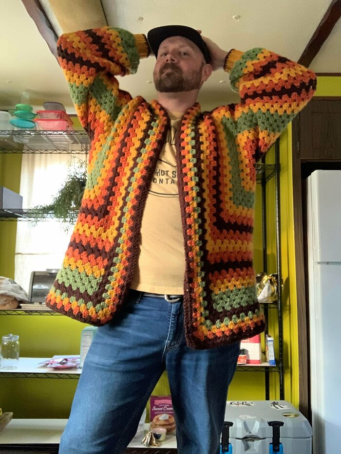 A man wears a brown, green, and orange striped crochet cardigan, a useful DIY creation.