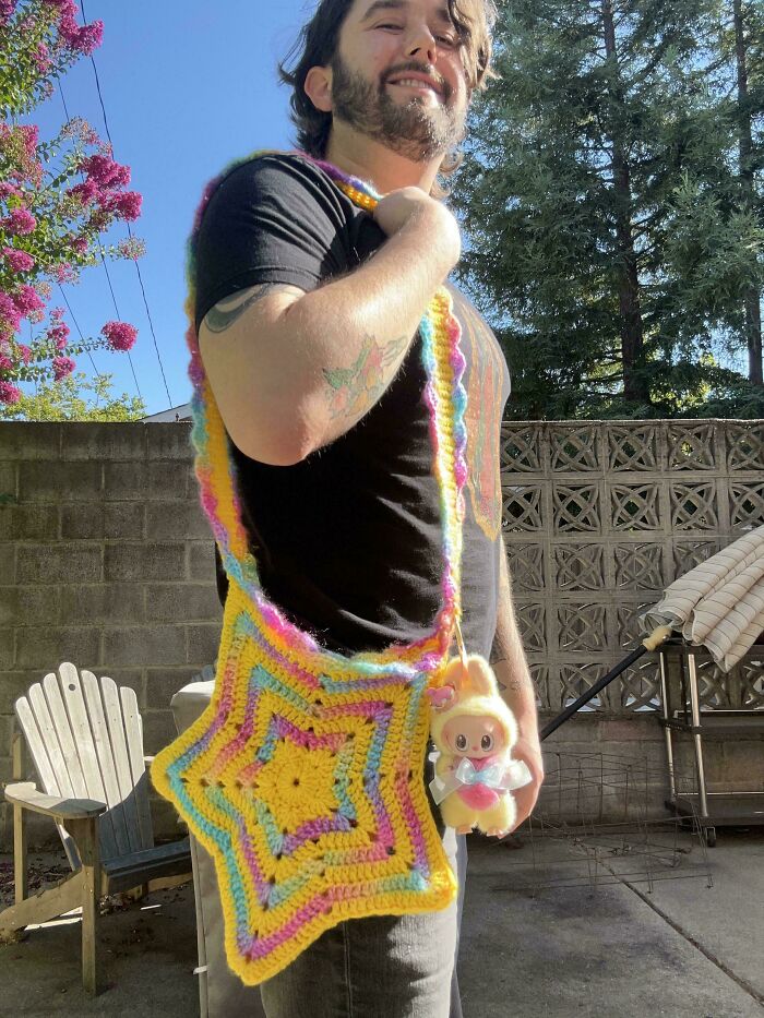 A man smiles, wearing a yellow star-shaped crochet bag with colorful edges and a small toy, a useful DIY creation.