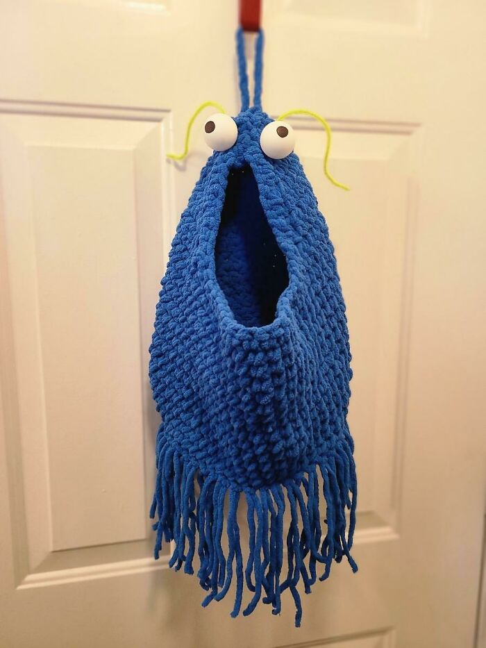 A fluffy blue crochet monster bag with googly eyes and yellow antennae hanging on a white door, a useful DIY creation.